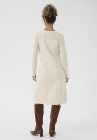 Térdig érő krémszínű kötött pulóverruha textúrával és hosszú ujjakkal, barna velúr magassarkú csizmával párosítva. Minimalista dizájn, látható díszítőelemek nélkül.