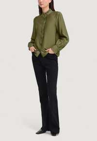 Blusa verde oliva con un colletto alto e dettagli a ruffles sul davanti. Maniche lunghe e voluminose, polsini con bottoni, abbinata a pantaloni neri a zampa d'elefante.