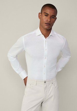 Camisa blanca de manga larga y botón, hecha de una tela ligera, con un cuello clásico y mangas con puños, combinada con pantalones de un color claro.