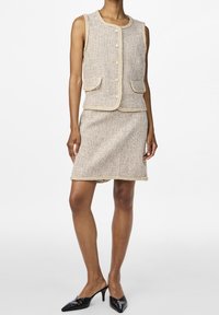 Beżowa kamizelka z tkaniny tweedowej, z okrągłym dekoltem, z przodu zapinana na guziki i z dwiema kieszeniami, zestawiona z dopasowaną spódnicą do kolan, wykończoną kontrastowym obszyciem.