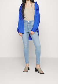 Blå oversized skjorta, beige linne, ljusblå skinny jeans och olivgröna klackskor med öppen tå. Släta texturer genomgående.