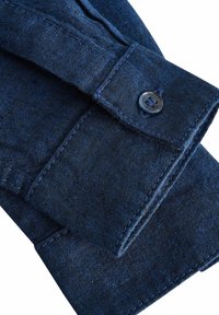 Polsino di una camicia in denim blu scuro con bottone, caratterizzato da una texture liscia e dettagli di cucitura ben visibili. Il tessuto ha una leggera lucentezza.