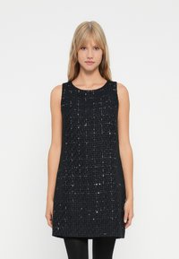 Jeune femme aux cheveux blonds portant une robe mini noire sans manches en texture avec des détails brillants subtils.