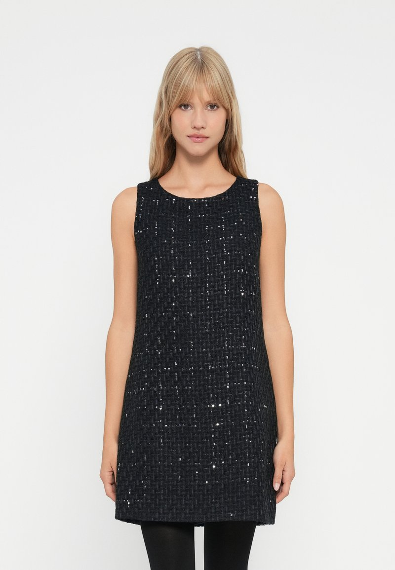 Jeune femme aux cheveux blonds portant une robe mini noire sans manches en texture avec des détails brillants subtils.