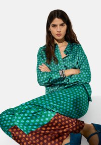 Zielona bluzka w kropki o luźnym kroju, zestawiona z spódnicą w niebiesko-brązowe wzory. Gładki materiał z delikatnym połyskiem.