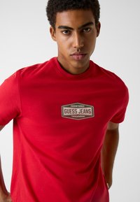 Czerwona bawełniana koszulka z prostokątną etykietą "GUESS JEANS" w ciemnobrązowym kolorze na środku, krótkimi rękawami i okrągłym dekoltem.