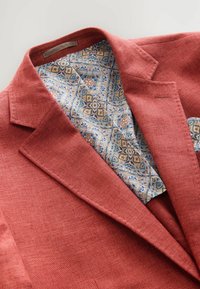 Rustfarbener Blazer aus strukturierter Stoff, mit einem gemusterten Innenfutter in Blau und Gold sowie einem kleinen Detail in Form eines Taschentuchs.