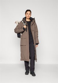 Long, brown insulated parka with a hood and large front pockets, worn over a black top and dark pants. Backpack with light-colored fabric and straps.