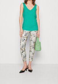 Tyrkysový pletený top bez rukávů s V-výstřihem, v kombinaci se vzorovanými slim-fit kalhotami s květinovými motivy. Drží zelenou kabelku a má na sobě černé podpatky.