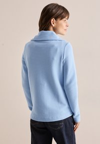 Pullover azzurro chiaro, strutturato, con colletto ampio, maniche lunghe e orlo dritto. Abbinato a jeans scuri, che mostrano uno stile casual.