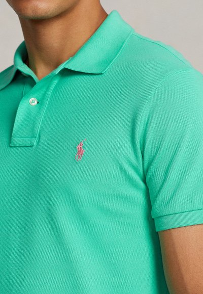 Πράσινο πόλο μπλουζάκι από υφασμάτινη υφή, με γιακά και μικρό ροζ λογότυπο στο αριστερό στήθος. Κοντά μανίκια με ριμπ μανσέτες.