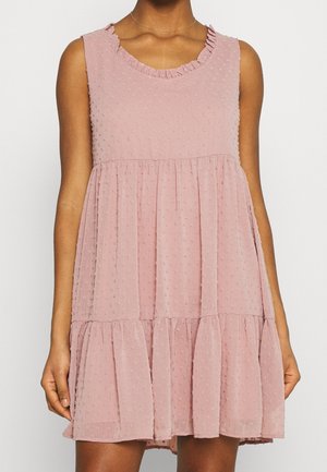 Vestido sin mangas de largo hasta la rodilla en color rosa claro con puntos texturizados, escote con volantes y falda de capas, vestido por una persona de piel morena.