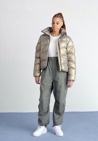 Giacca puffer metallic oro con collo alto, abbinata a pantaloni cargo verde oliva. Il modello indossa una maglietta bianca e sneakers bianche.