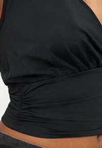 Κοντινό πλάνο μαύρου αμάνικου τοπ με πτυχωτές πλευρές και εφαρμοστό ζωνάκι στη μέση, πάνω στον κορμό ενός ατόμου.