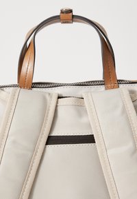Sac en toile beige avec des poignées en cuir marron. Comprend une fermeture éclair et une poche interne avec une fermeture éclair foncée. Coutures durables visibles.