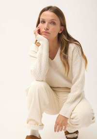 Cremfarbener Strickpullover mit V-Ausschnitt und gerippter Textur, kombiniert mit passenden weit geschnittenen gerippten Hosen. Das Outfit hat umgeschlagene Ärmel.