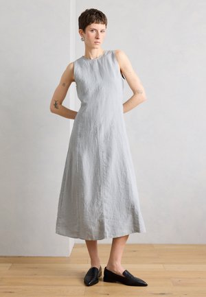 Person mit kurzen Haaren, die ein ärmelloses graues Midikleid und schwarze Slip-On-Schuhe trägt, steht auf einem Holzboden vor einer hellen Wand.