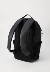 Sac à dos noir avec des bretelles rembourrées et texturées, des poches latérales et un extérieur lisse. Il dispose d'une poignée supérieure pour le transport.