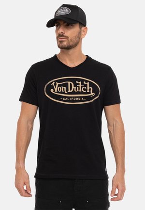 Homme portant un t-shirt noir Von Dutch California et une casquette noire assortie Von Dutch, regardant sur le côté sur un fond uni.