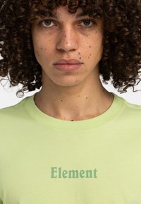 Světle zelené tričko s crew neck výstřihem s nápisem "Element" v tmavě zeleném písmu. Materiál vypadá měkce, s hladkou, nezdobenou texturou.