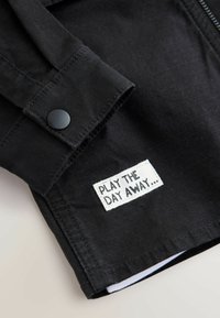 Črna tkanina jakna z zapenjanjem na gumb in belim pravokotnim obližem s črnim napisom "IGRAJ DAN DO KONCA." Teksturiran zaključek.