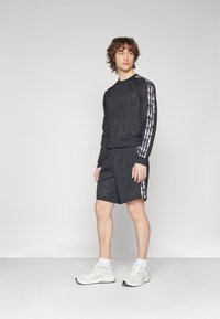 Crni mrežasti top s dugim rukavima i prugastim detaljima, uparen s crnim šortsevima. Model nosi bijele atletske tenisice i čarape do gležnjeva. Minimalistički dizajn.