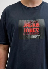 Navy blå bomulds t-shirt med korte ærmer. Har et fremtrædende sort og rødt grafisk design med teksten "JACK & JONES" i fed typografi.