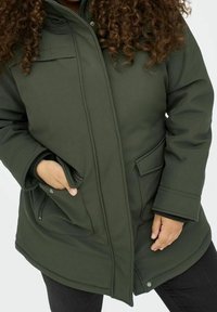 Mörkgrön, vadderad parka-jacka med hög krage, dragkedjefickor och breda ärmslut. Slät textur med en åtsittande silhuett och framknäppning.