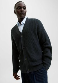 Cardigan tricoté gris foncé avec un col en V, arborant un motif texturé et quatre boutons. Porté par-dessus une chemise à col bleu clair, associé à un pantalon rayé.