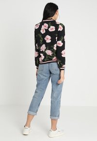 Giacca bomber floreale con sfondo nero, caratterizzata da grandi fiori rosa e foglie verdi. Abbinata a jeans azzurri arrotolati e sneakers bianche.