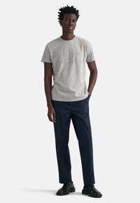 T-shirt en coton gris avec logo en relief, associée à un pantalon bleu marine et des sandales noires. Le modèle se tient les mains dans les poches, devant un fond neutre.
