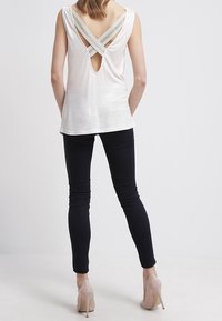 Top blanco sin mangas con diseño de espalda entrelazada y un pequeño corte. Combinado con pantalones ajustados negros y tacones nude.