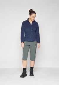 Navy fleece zip jacket with a stand collar, paired with grey capri pants and black hiking boots. Soft texture, functional design.