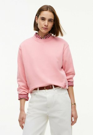 Kvinde med kort brunt hår iført pink sweatshirt over rød- og hvide stribet skjorte og hvide bukser med brun bælte mod hvid baggrund.