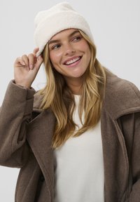 Biely, nadýchaný čiapka s prehnutým okrajom, nosená s hnedým vlneným kabátom cez bielu blúzku. Tkanina vyzerá mäkko a textúrovane.