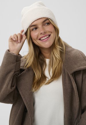 Weißer, flauschiger Beanie mit umgeschlagenem Rand, getragen zu einem braunen Wollmantel über einem weißen Oberteil. Der Stoff wirkt weich und strukturiert.