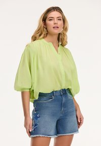 Blusa verde chiaro, trasparente, a maniche corte con colletto alla cinese e chiusura a bottoni, abbinata a pantaloni corti in denim blu con ricami floreali.