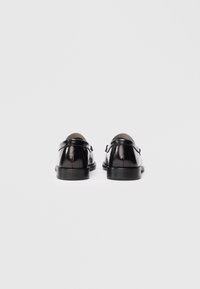 Um par de sapatos de vestir em couro preto mostrados de trás sobre um fundo branco liso, destacando seu acabamento polido e saltos baixos.