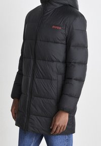 Schwarze Steppjacke mit Kapuze, die eine horizontale gesteppte Struktur und ein rotes Logo auf der linken Brustseite aufweist. Mit Reißverschluss an der Vorderseite und seitlichen Taschen.