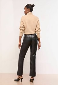 Sieviete stāv ar muguru, ģērbusies bēša džemperī ar augstu apkakli, melnās ādas biksēs un melnās papēžu siksnu kurpēs.