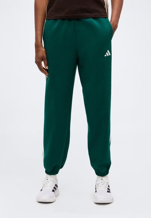 Person trägt dunkelgrüne Adidas-Sweatpants mit elastischen Bündchen, weiße Socken und weiße Adidas-Sneaker und steht mit den Händen in den Taschen.