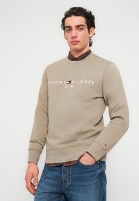 Μπεζ φούτερ με στρογγυλή λαιμόκοψη, με λευκό κείμενο "TOMMY HILFIGER EST. 1985". Φορεμένο πάνω από πουκάμισο μπορντό καρό, με λεπτομέρειες λογότυπου στο μανίκι.