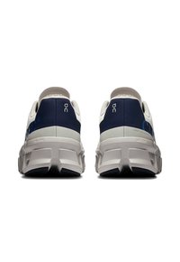 Des chaussures de sport blanches et bleu marine avec une texture lisse, dotées d'un col rembourré, d'un logo au niveau du talon et d'une semelle épaisse et segmentée.