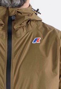 Brązowa lekka kurtka z kapturem, z czarnym zamkiem błyskawicznym i wielokolorowym logo na lewej piersi. Gładki, wodoodporny materiał.
