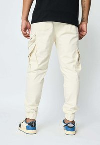 Pantalons cargo beiges avec des poignets de cheville élastiques, plusieurs poches latérales et une coupe décontractée, associés à des baskets bleues et noires.