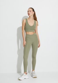 Sujetador deportivo verde oliva con tirantes anchos, combinado con leggings de cintura alta que presentan un diseño de cintura cruzada. El modelo lleva zapatillas blancas.