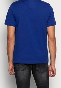 Hombre con una camiseta de manga corta azul lisa y jeans negros desgastados, visto desde atrás contra un fondo liso.