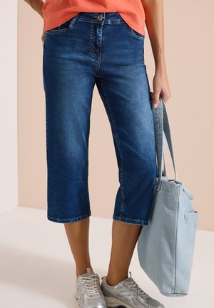 Vrouw die blauwe denim capribroek draagt, witte sneakers en een lichtblauwe draagtas vasthoudt tegen een beige achtergrond.