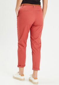Pantalon en coton rouge à coupe fuselée avec des revers, doté de poches arrière et d'une ceinture tressée marron. Porté avec des baskets blanches.