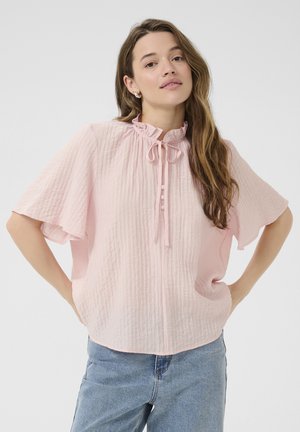 Blouse rose pâle en tissu texturé, avec un col froncé, un détail à nouer et des manches courtes. Boutons placés de manière décorative à l'avant.